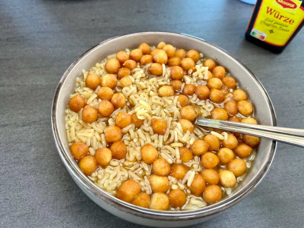Eine Schüssel mit dampfender Buchstabensuppe, die reichhaltig mit goldbraun gebackenen Backerbsen garniert ist. Ein silberner Löffel steckt in der Suppe. Im Hintergrund steht eine Flasche Maggi-Würze auf dem grauen Tisch des Wohnmobils.