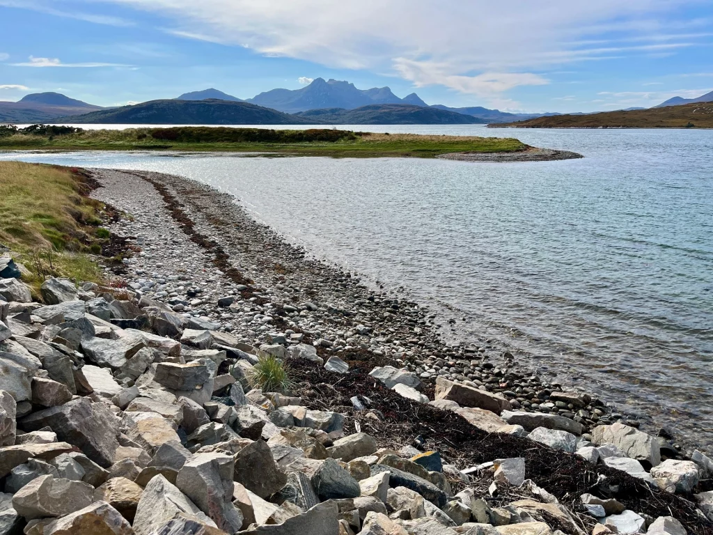 Ein steiniges Ufer an einem ruhigen Meeresarm, im Hintergrund die Silhouette der schottischen Berge. 