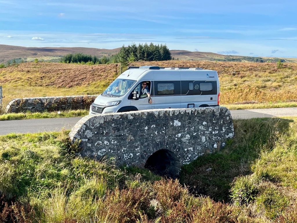 Ein silberner Kastenwagen-Camper ('Kätter') fährt über eine kleine, urige Steinbrücke mit rundem Durchlass in der Nähe von Melvich. Die Brücke ist von wildem, herbstlichem Gras und Heidekraut umgeben, im Hintergrund erstrecken sich die sanften Hügel von Sutherland unter blauem Himmel.