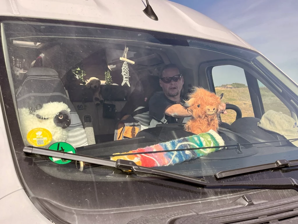 Perspektive durch die Windschutzscheibe des Campers: Ein Fahrer am Lenkrad, auf dem Armaturenbrett sitzen kleine Reisebegleiter (Kuscheltiere). Der Blick geht hinaus auf die weite, sonnige Highland-Landschaft.