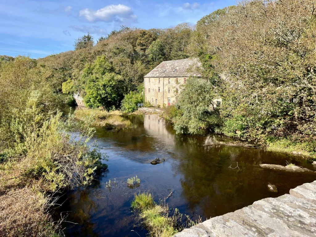 Ein altes Steinhaus, das direkt an einem Flussufer liegt und von dichtem Gebüsch und Bäumen eingerahmt wird – ein typisches Beispiel für die historische Architektur in der Region Sutherland.