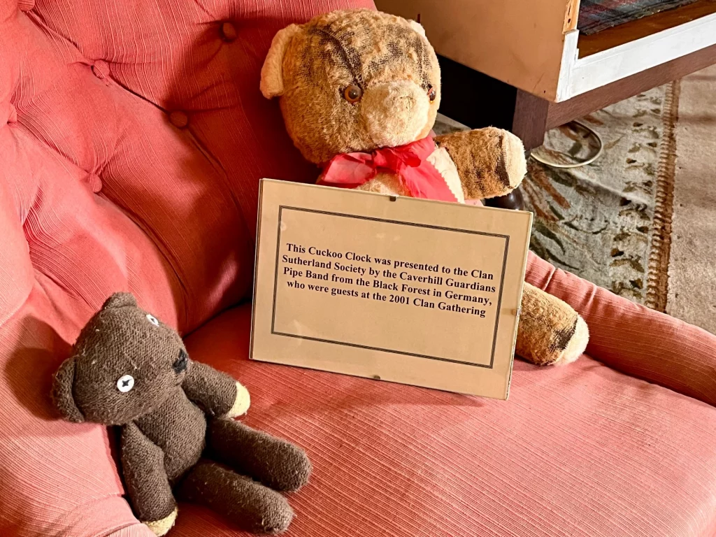 Zwei Teddybären auf einem rosa Sessel neben einer gerahmten Urkunde über das Geschenk der Kuckucksuhr aus dem Jahr 2001.