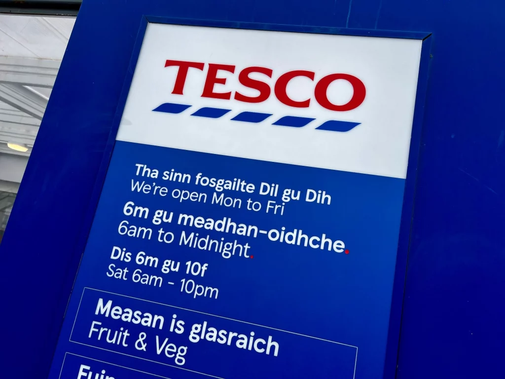 Ein blaues Hinweisschild des Supermarktes Tesco in Stornoway, Schottland. Die Öffnungszeiten und Abteilungen wie 'Fruit & Veg' sind sowohl auf Englisch als auch auf Gälisch beschriftet.