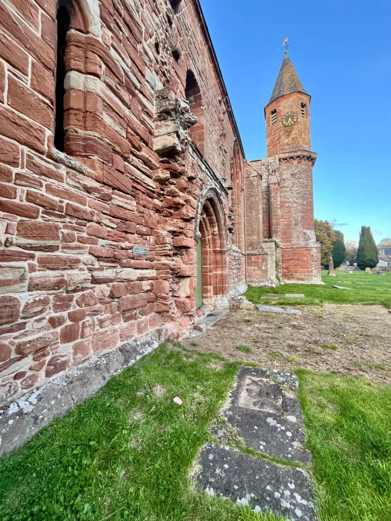 Seitenansicht der Außenmauer aus rotem Sandstein mit Blick auf den erhaltenen Treppenturm mit Uhr und Wetterfahne.