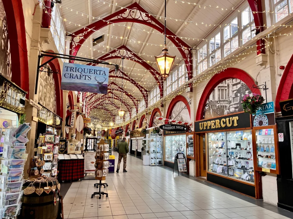 Innenansicht einer viktorianischen Einkaufspassage mit hohen Decken, dekorativen roten Holzbögen und zahlreichen kleinen Fachgeschäften wie "Triquerta Crafts" und "Uppercut". Lichterketten überspannen den Gang.