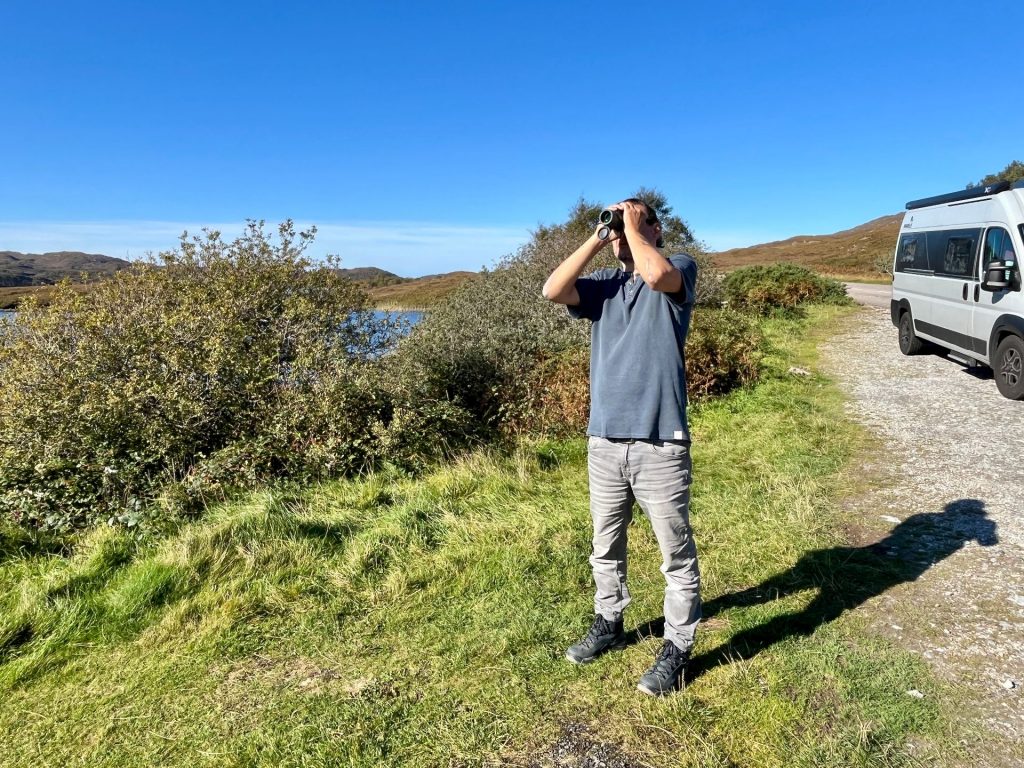 Ein Mann steht auf einer Wiese neben einer Straße in den schottischen Highlands und schaut durch ein Fernglas über die Landschaft.