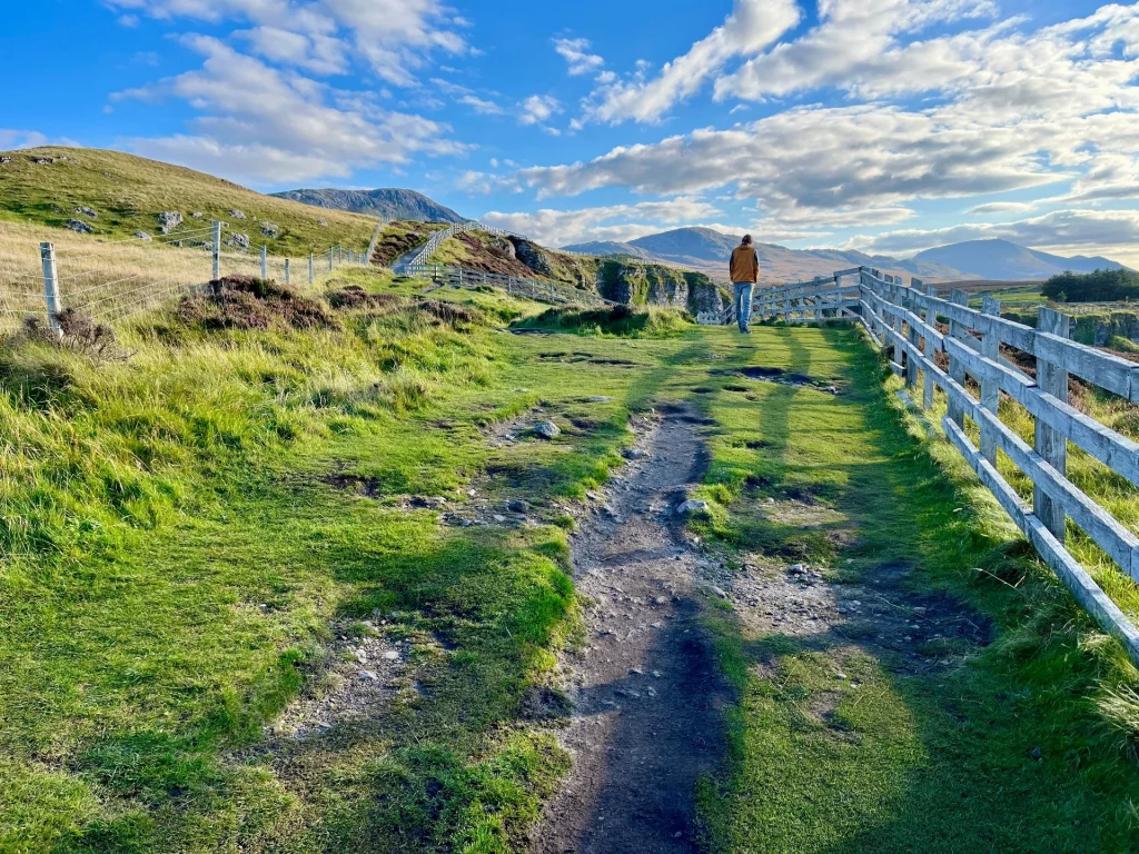 Ein Wanderer von hinten gesehen auf einem Pfad, der von einem rustikalen Holzzaun flankiert wird. Im Hintergrund erheben sich majestätische Berge unter einem Wolkenhimmel.