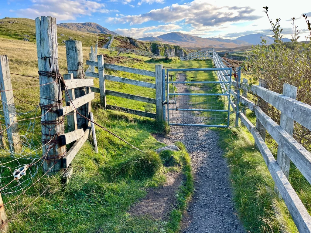 Ein rustikales Metallgatter unterbricht einen Schotterpfad, der durch grüne Weiden führt. Alte Holzpfosten mit Stacheldraht säumen den Weg in Richtung der fernen Berge.