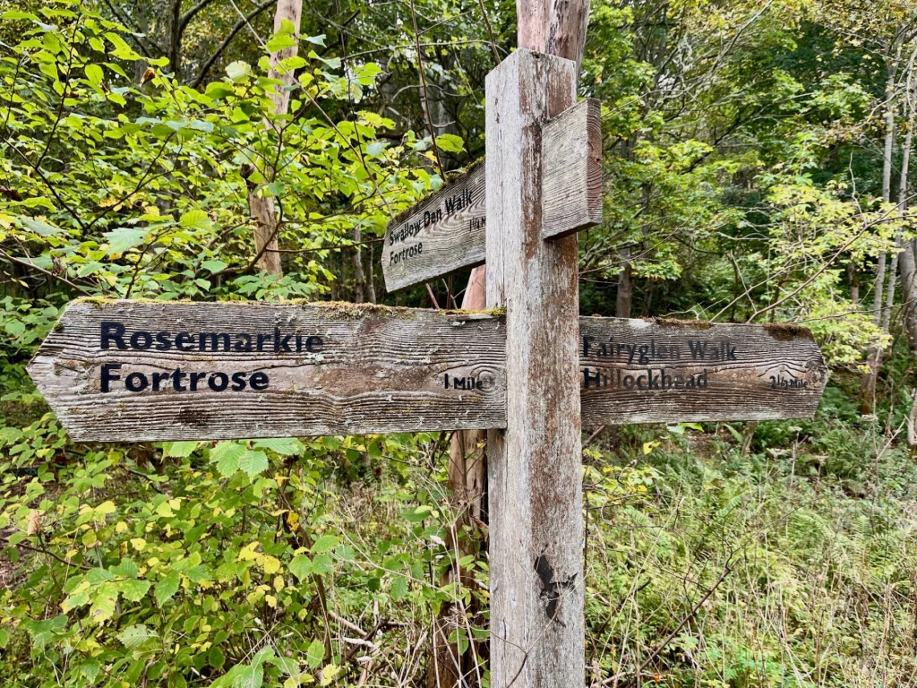 Ein hölzerner Wegweiser im Wald, der die Richtungen zum Fairy Glen Walk, Rosemarkie und Fortrose anzeigt.