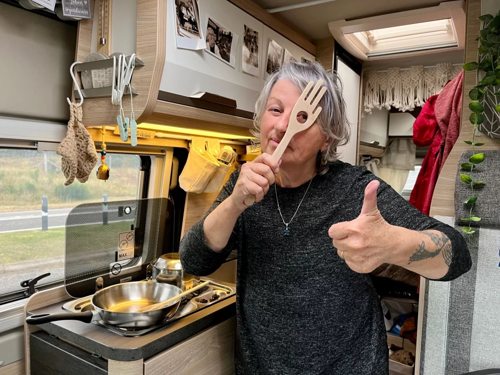 Karin steht lächelnd in einer Wohnmobil-Küche, hält sich scherzhaft eine hölzerne Kochgabel vor das Auge und gibt einen Daumen nach oben.