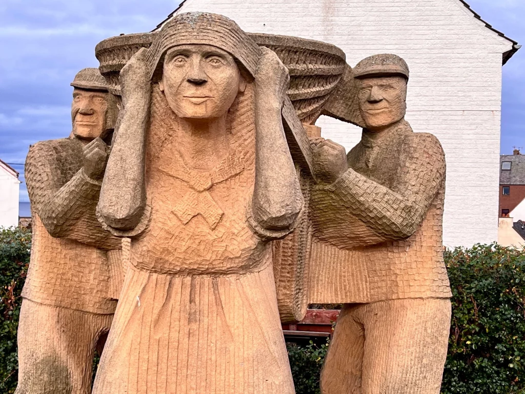 Nahaufnahme der Sandsteinskulptur „The Creel Loaders“ in Dunbar. Sie zeigt eine Frau mit einem großen geflochtenen Fischkorb auf dem Rücken, flankiert von zwei Männern, die ihr beim Tragen der schweren Last helfen. Die Gesichter und Kleider sind im groben, ausdrucksstarken Stil des lokalen Sandsteins gehauen.