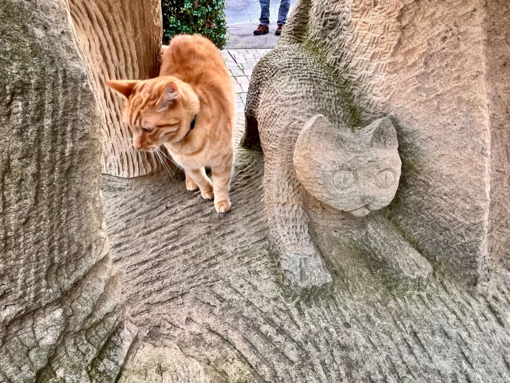 Detailaufnahme am Fuß der Creel-Loaders-Skulptur. Eine echte, rot-getigerte Katze läuft direkt neben einer in den Stein gehauenen Katze über den Sockel. Der Kontrast zwischen der lebendigen Katze und der steinernen Figur wirkt wie ein gewollter, mystischer Moment.
