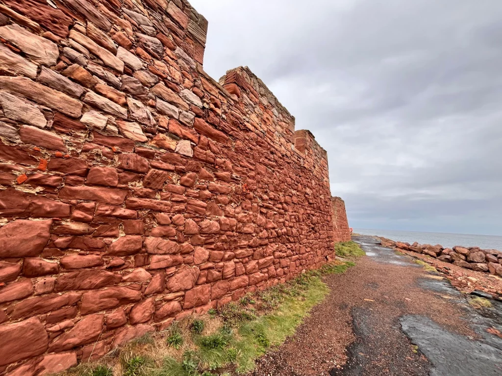 Eine lange, hohe Mauer aus rotem Sandstein führt entlang eines Weges direkt am Meer.