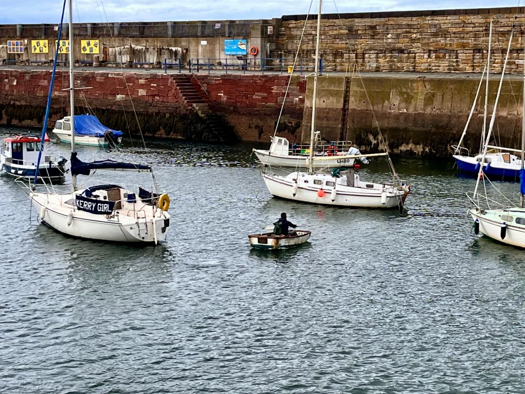 Verschiedene kleine Segelboote und Fischerboote ankern im ruhigen Wasser des Hafens vor einer hohen Steinmauer.