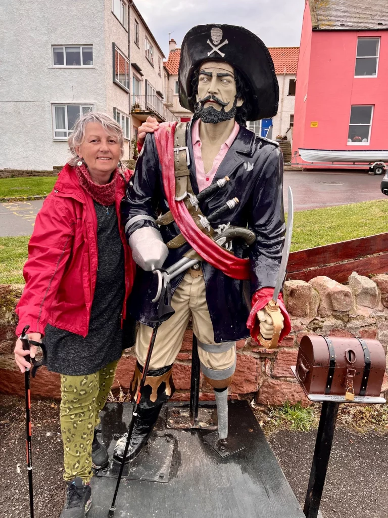 Karin steht in roter Jacke lächelnd neben einer lebensgroßen Piratenfigur mit Holzbein am Hafen von Dunbar.