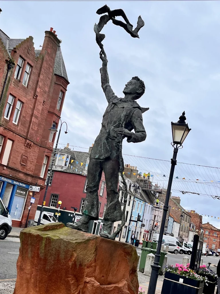 Blick auf die Bronzeskulptur des jungen John Muir auf der High Street in Dunbar, Schottland. Die Statue steht auf einem Sockel aus rotem Sandstein. Muir ist in Wanderkleidung dargestellt und blickt nach oben, während sein rechter Arm ausgestreckt ist und Vögel in den Himmel entlässt. Im Hintergrund sind historische rote Sandsteingebäude, eine bunte Häuserzeile, eine Straßenlaterne und ein Gerüst zu sehen.
