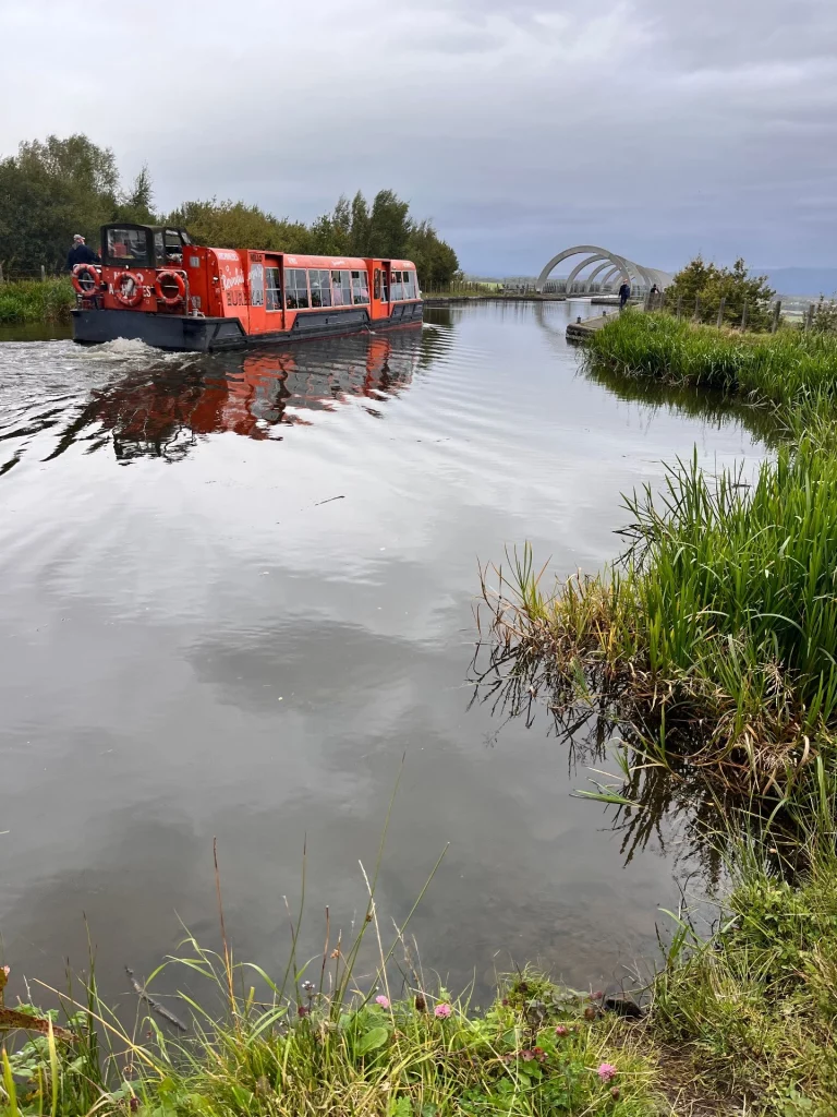Ein rotes Kanalboot fährt auf dem ruhigen Wasserweg in Richtung der Betonbögen des Falkirk Wheels, gesäumt von Schilf und Bäumen.