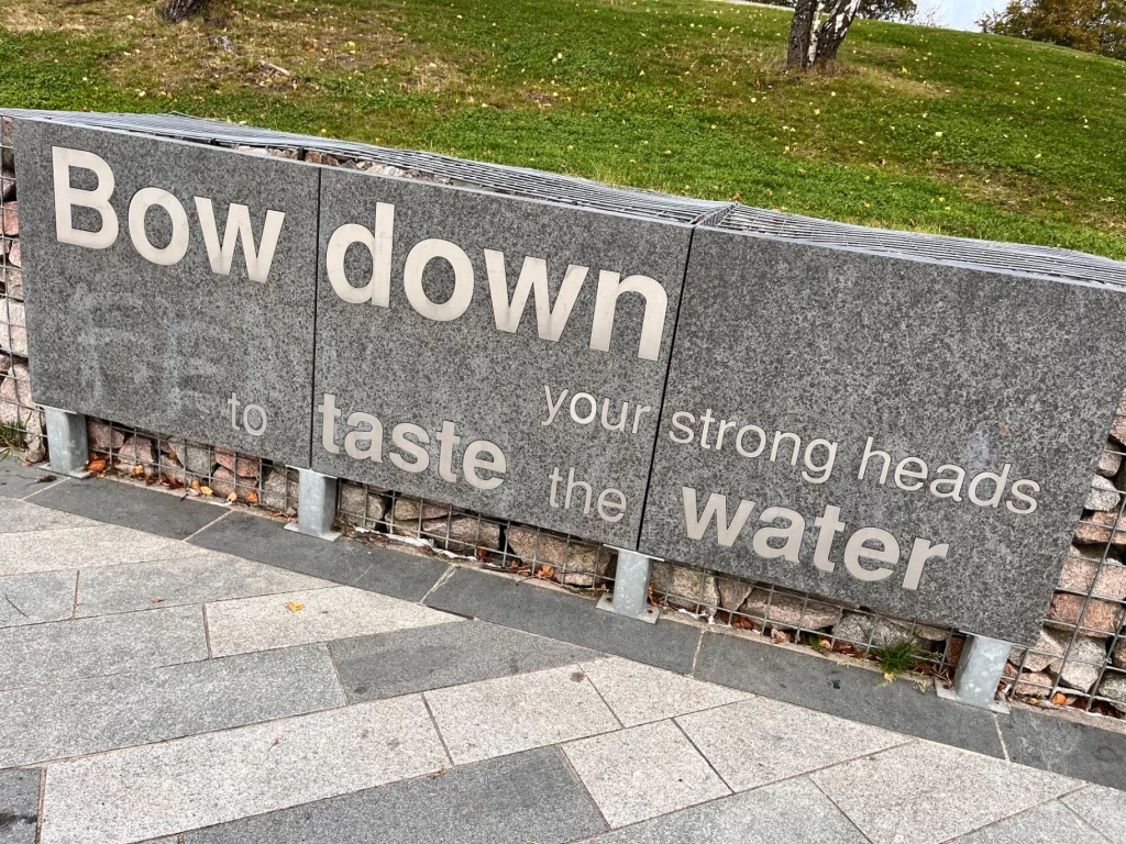 Eine graue Steinmauer mit der Inschrift "Bow down your strong heads to taste the water" – ein poetischer Bezug zur Haltung der Kelpie-Skulpturen.
