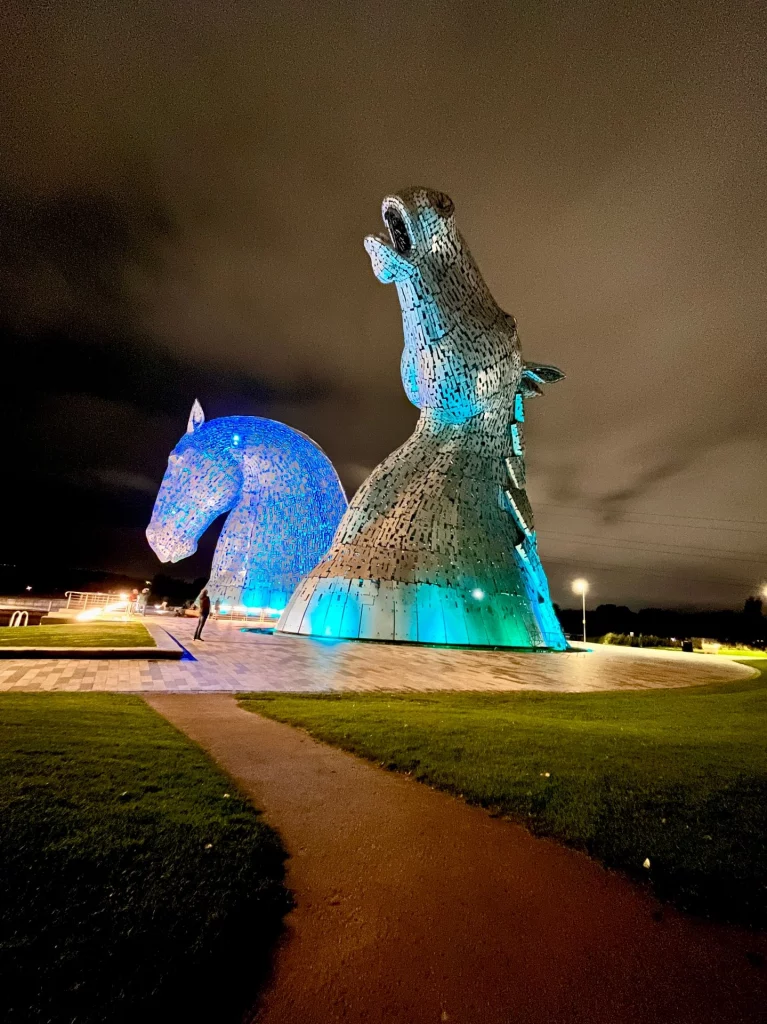 Seitenansicht der Kelpies-Skulpturen bei Nacht, die in leuchtendem Blau und Türkis angestrahlt werden, davor ein kleiner Pfad.