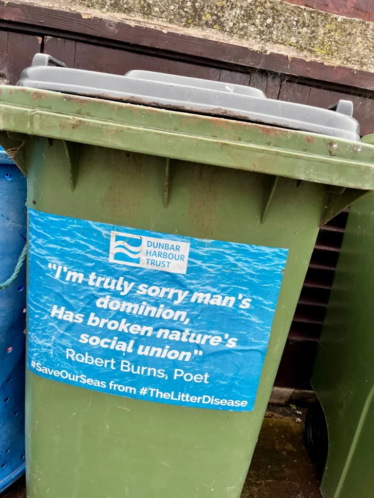 Eine grüne Mülltonne mit einem Aufkleber, der ein Zitat von Robert Burns trägt: „I'm truly sorry man's dominion, Has broken nature's social union.“
