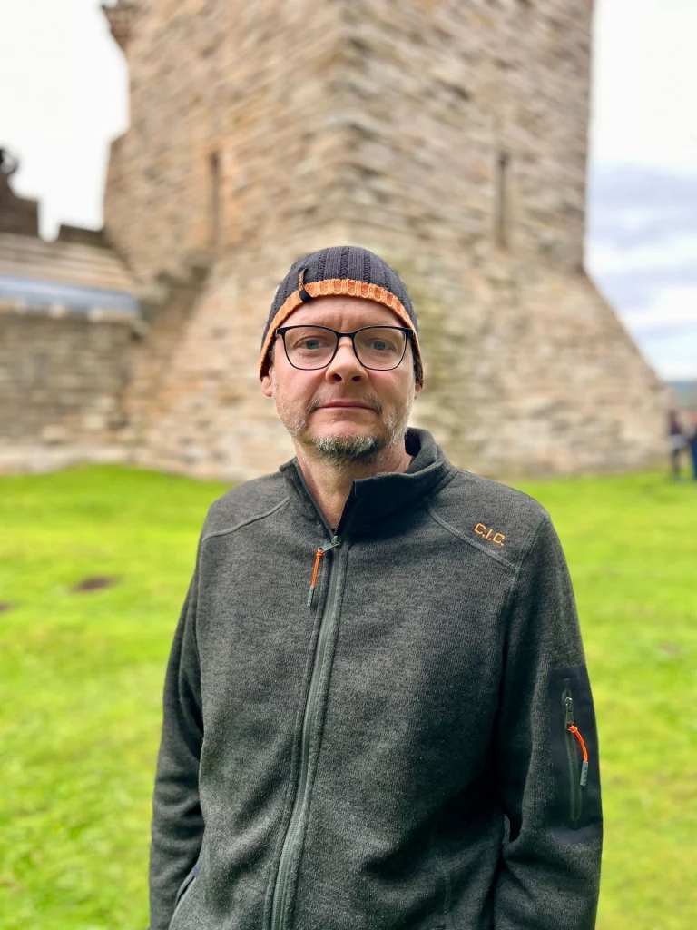 Porträtaufnahme eines Mannes mit Brille und Mütze vor dem unscharfen Hintergrund des Wallace Monument.