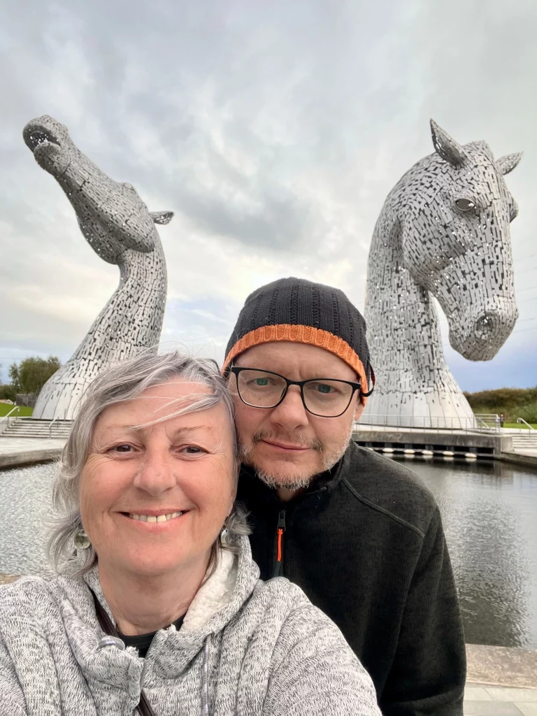 Ein lächelndes Paar macht ein Selfie im Helix Park in Falkirk, im Hintergrund ragen die beiden riesigen Kelpie-Pferdekopfskulpturen aus Stahl auf.