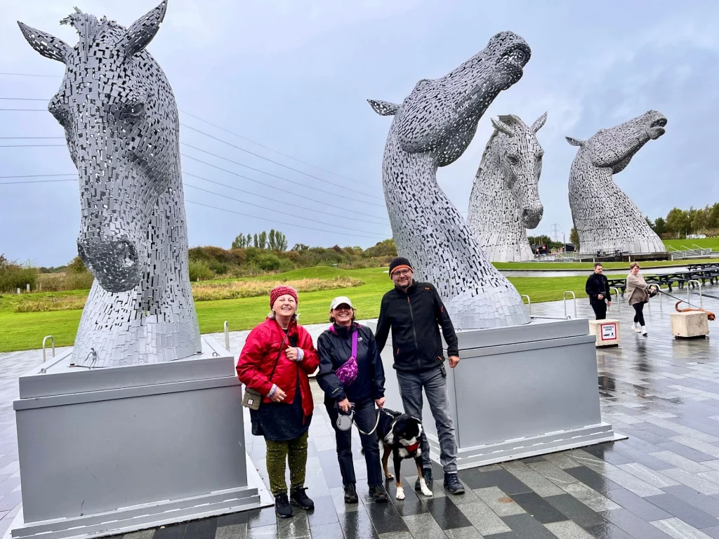 Drei lächelnde Personen, Ingo, Karin und Kathryn, stehen zusammen mit dem Hund Milo vor den imposanten Kelpies-Skulpturen in Schottland. Karin trägt eine leuchtend rote Jacke, Kathryn eine lila Bauchtasche und Ingo eine dunkle Jacke und Mütze. Sie posieren auf einem gepflasterten Platz zwischen den beiden riesigen Pferdekopf-Skulpturen aus Edelstahl. Der Himmel ist bewölkt.