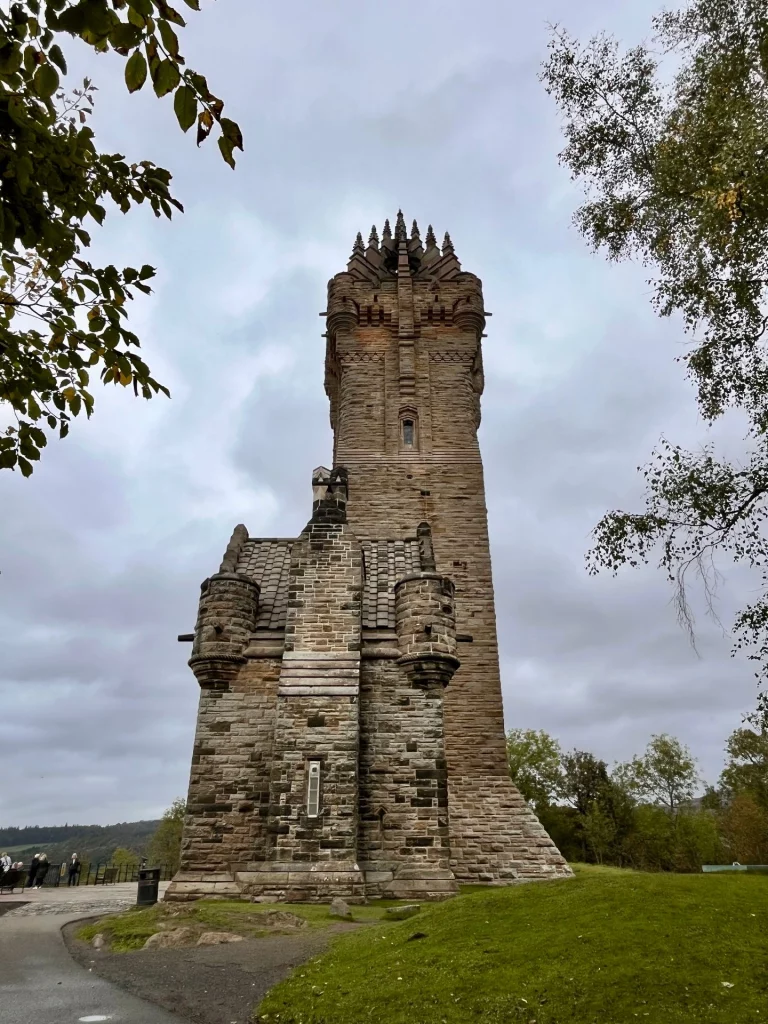 Seitenansicht des National Wallace Monument in Stirling unter bewölktem Himmel. Der massive, sandsteinfarbene Turm im viktorianisch-gotischen Stil ragt über ein kleineres Gebäude mit Ecktürmchen hinaus, umgeben von grünen Rasenflächen und herbstlichen Bäumen.
