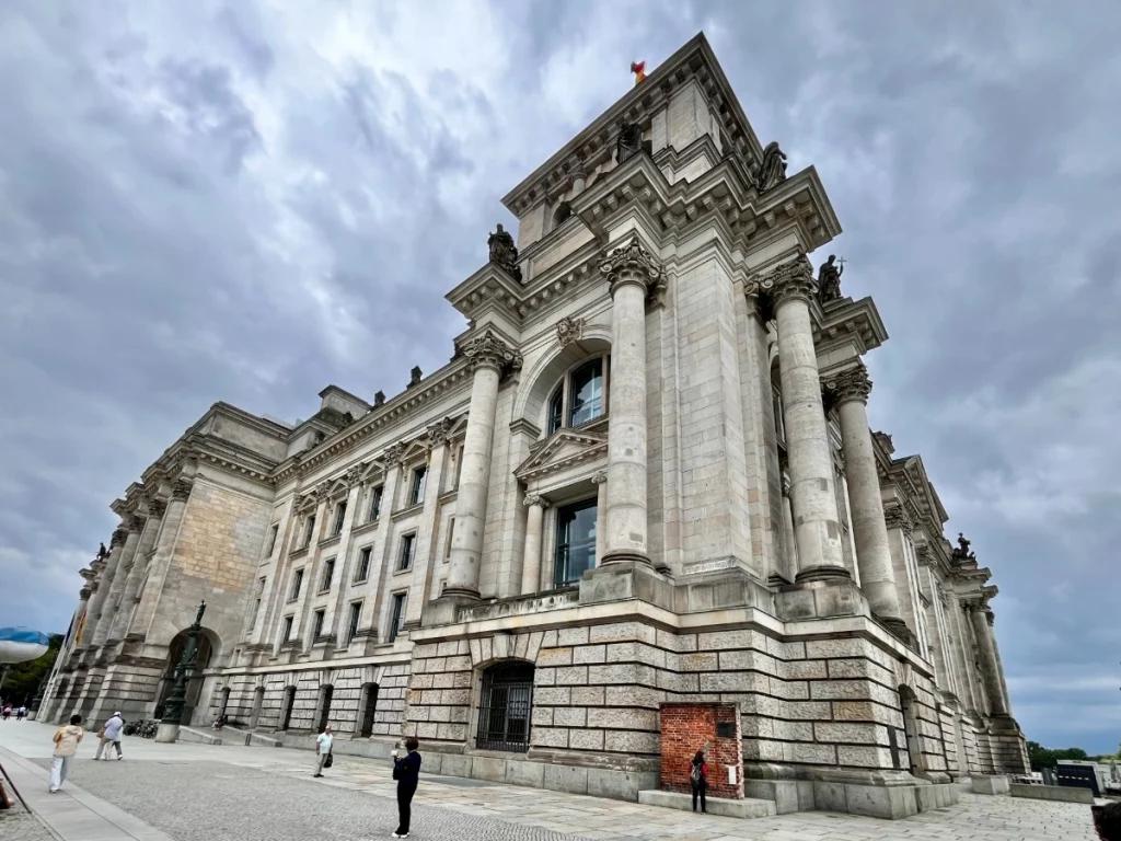 Die imposante Außenfassade des Reichstagsgebäudes in Berlin unter einem dramatischen, bewölkten Himmel. Die Aufnahme aus einer tiefen Perspektive betont die mächtigen korinthischen Säulen und die detailreichen Steinmetzarbeiten des historischen Bauwerks. Im Vordergrund ist der weitläufige Platz vor dem Parlamentssitz zu sehen.