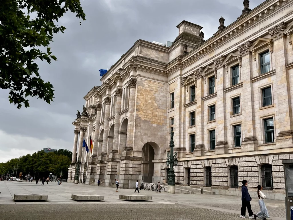 Die imposante Außenfassade des Reichstagsgebäudes in Berlin unter einem dramatischen, bewölkten Himmel. Die Aufnahme aus einer tiefen Perspektive betont die mächtigen korinthischen Säulen und die detailreichen Steinmetzarbeiten des historischen Bauwerks. Im Vordergrund ist der weitläufige Platz vor dem Parlamentssitz zu sehen.
