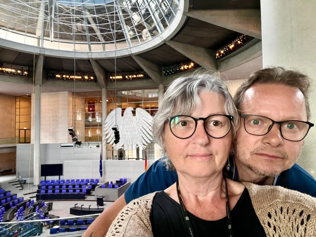 Ein Selfie-Porträt eines Paares im Plenarsaal des Deutschen Bundestages in Berlin. Im Hintergrund ist die markante Architektur des Saals mit den blauen Abgeordnetensitzen, dem großen Bundesadler und der modernen Glaskuppel-Konstruktion zu sehen.