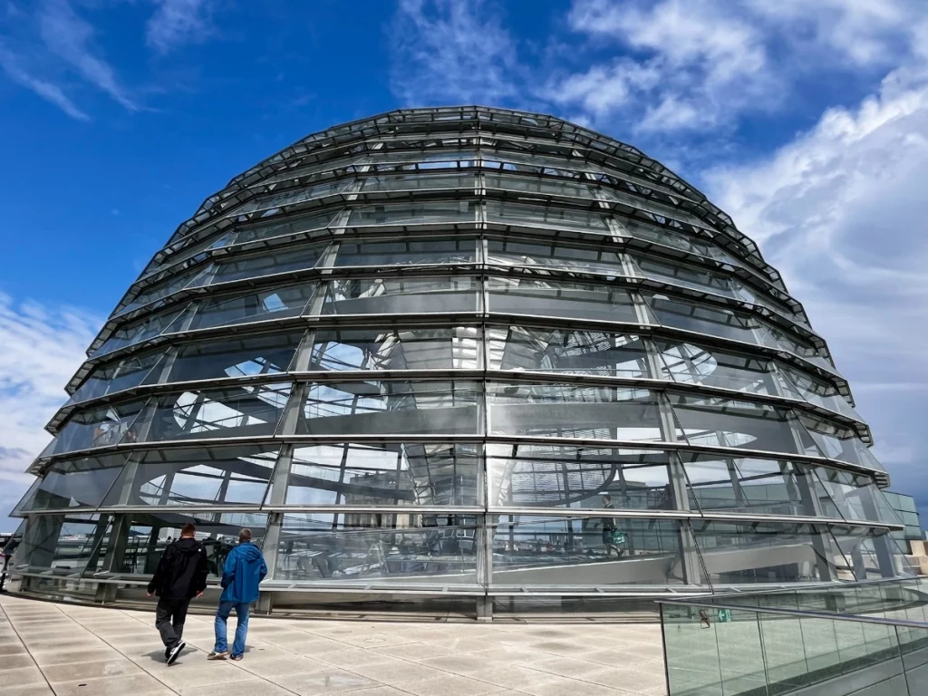 Eine Außenansicht der modernen Glaskuppel auf dem Dach des Reichstagsgebäudes unter einem strahlend blauen Himmel mit lockeren Wolken. Die halbrunde Konstruktion aus Glas und Stahl lässt tief in das Innere blicken, wo das trichterförmige Spiegelsystem zu erkennen ist. Im Vordergrund spazieren zwei Besucher über die weitläufige Dachterrasse in Richtung der Kuppel.