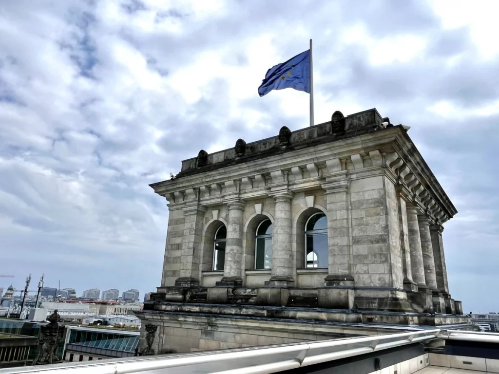 Ein Zeichen für Europa über den Dächern Berlins: Die europäische Flagge weht stolz auf einem der historischen Türme des Reichstags. Dieser Anblick verbindet die beständige Geschichte des Gebäudes mit der modernen, gemeinschaftlichen Zukunft. Ein kurzer Blick über die Brüstung der Dachterrasse zeigt zudem, wie weitläufig sich die Hauptstadt von hier oben erstreckt.