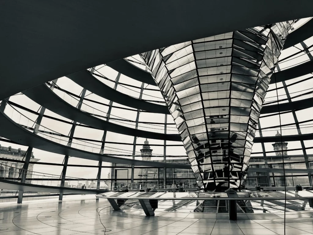 Spiel mit Licht und Form: Diese Schwarz-Weiß-Ansicht betont die futuristische Eleganz der Kuppelarchitektur. Die Spiegelungen auf dem zentralen Trichter und die klare Linienführung der Rampen erzeugen eine fast hypnotische Symmetrie. In der Reduktion auf Graustufen kommen die technischen Details und die beeindruckende Weite der Konstruktion von Norman Foster noch stärker zur Geltung.