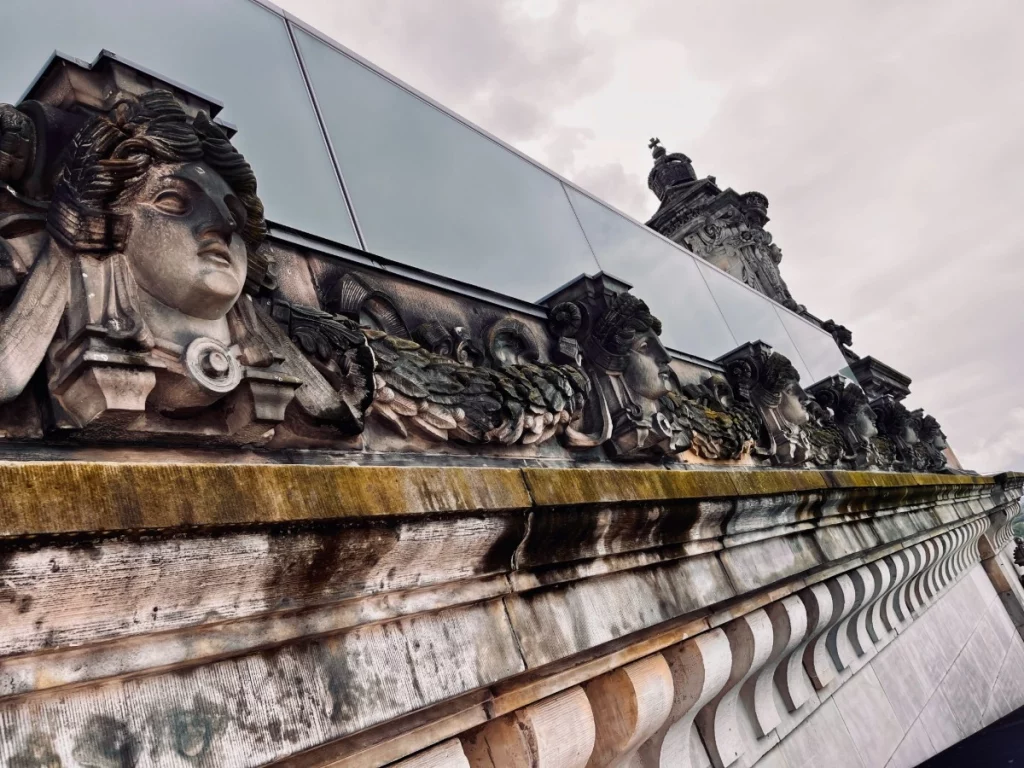 Detailreiche Geschichte trifft auf minimalistische Moderne: Während die historischen Steinmetzarbeiten an der Fassade des Reichstags von vergangener Pracht erzählen, spiegelt sich in der darüber liegenden Glasbrüstung der heutige Geist der Transparenz wider. Die Verwitterungsspuren auf dem Sandstein verleihen dem Gebäude eine authentische Patina und machen die bewegte Historie Berlins greifbar.