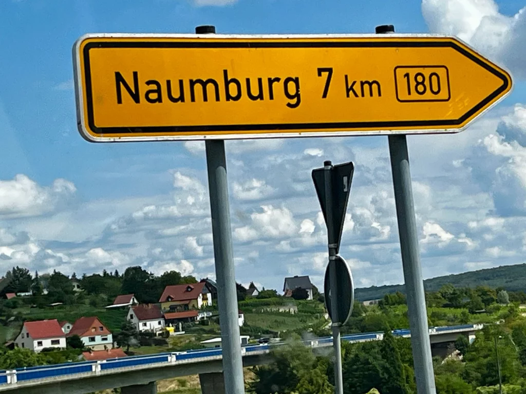 Ein gelbes Verkehrsschild weist nach rechts: 'Naumburg 7 km' an der Bundesstraße 180. Im Hintergrund eine hügelige Landschaft mit Häusern, Bäumen und einer Brücke unter blauem Himmel.