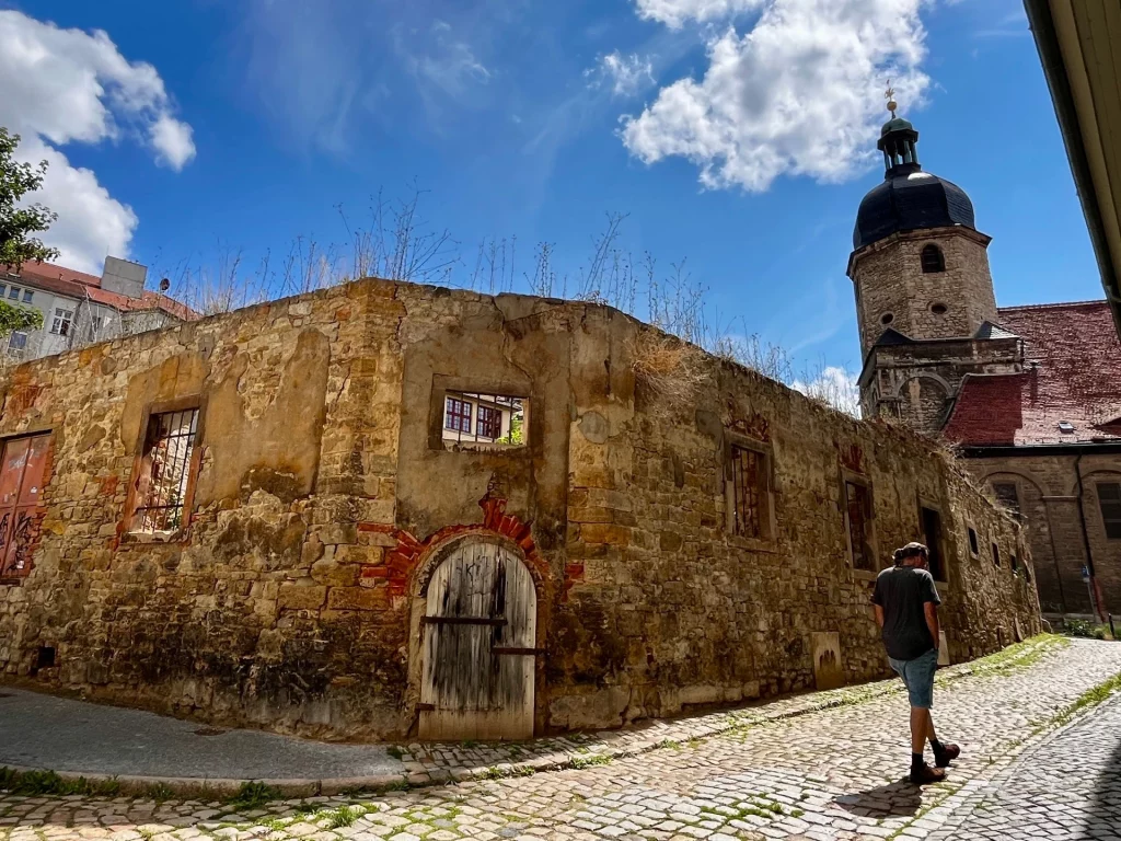 Natursteinruine ohne Dach mit Grasbewuchs auf den Mauerkronen, im Hintergrund ragt der massive Turm der Wenzelskirche mit barocker Haube in den blauen Himmel.