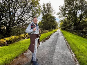Ein lächelnder Mann (Ingo) mit Brille, Rucksack und einer Flagge auf dem Hoodie steht auf einer nassen Landstraße, die von moosigen Steinmauern gesäumt ist.