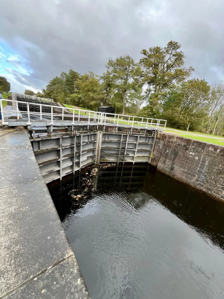 Blick auf die massiven Metall-Schleusentore und eine weiße Drehbrücke, die den Caledonian Canal überspannt, mit einem typischen weißen Highland-Haus im Hintergrund.