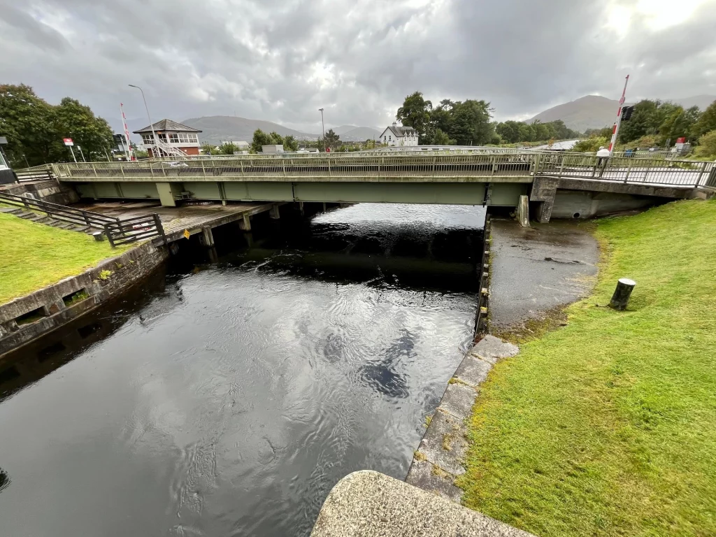 Eine Weitwinkelaufnahme der Banavie-Drehbrücke am Kaledonischen Kanal in Schottland unter einem bewölkten Himmel. Die grüne Metallbrücke führt über dunkles Wasser, das zwischen grasbewachsenen Ufern und Steinmauern fließt. Im Hintergrund sind Berge, ein kleines weißes Haus und ein Kontrollturm zu sehen.