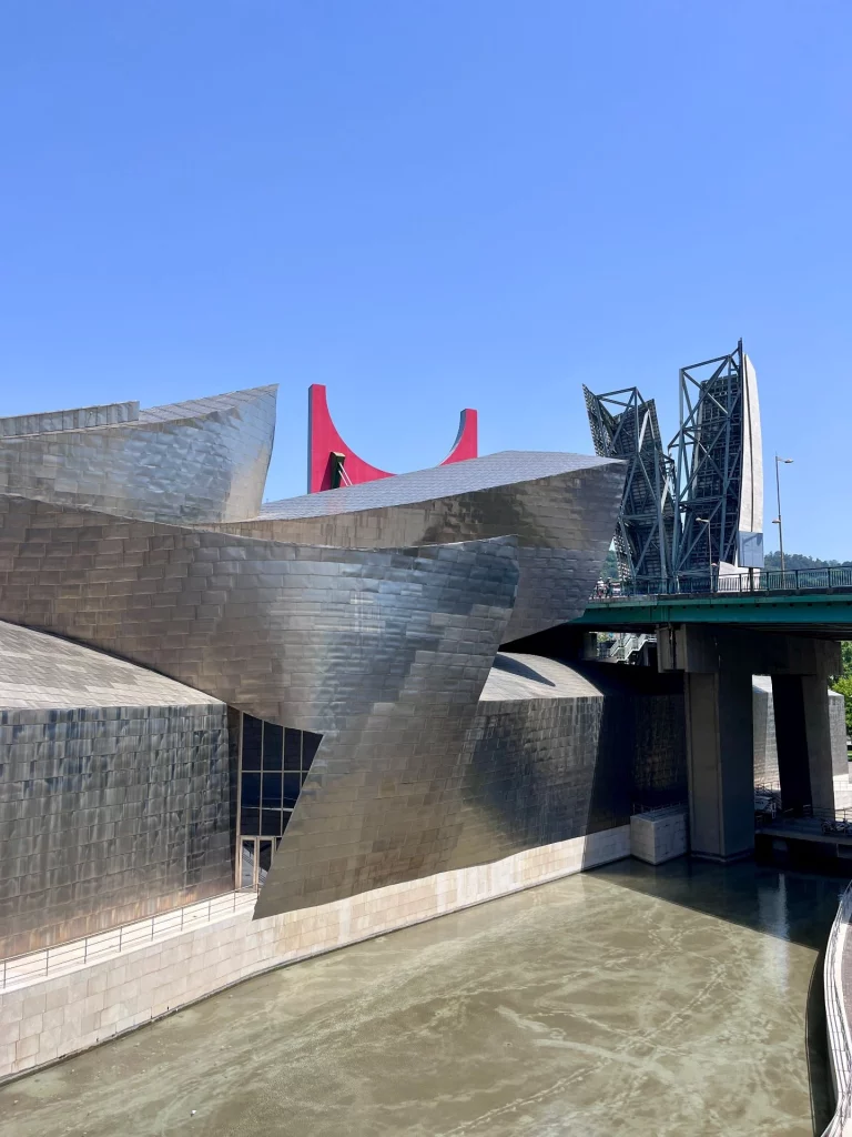 Außenansicht der geschwungenen Metallwände des Guggenheim-Museums in Bilbao, im Hintergrund die grüne La-Salve-Brücke mit dem roten Bogen-Kunstwerk von Daniel Buren.