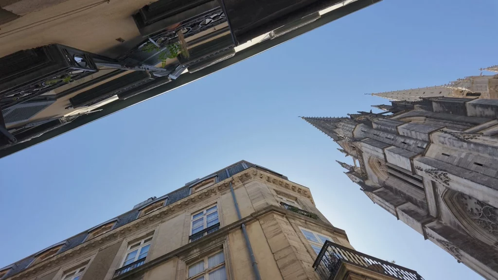 Blick aus der Froschperspektive nach oben zwischen historischen Altstadthäusern und der hoch aufragenden Turmspitze der Kathedrale von Bayonne gegen den blauen Himmel.