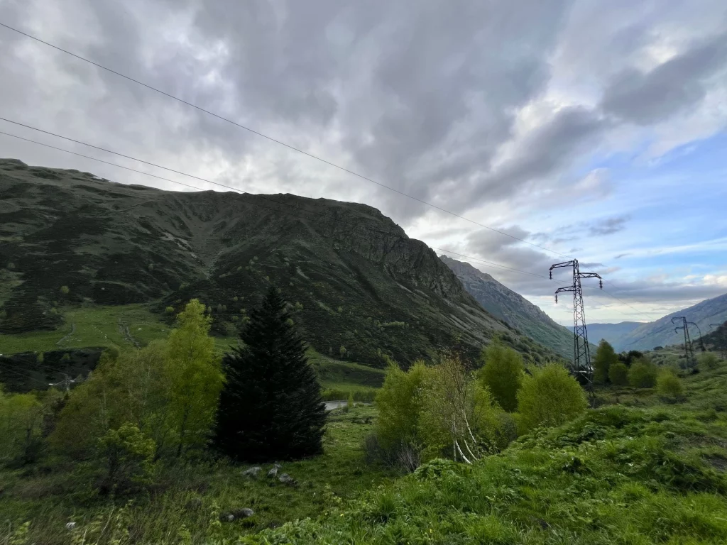 Eine grüne Berglandschaft in den Pyrenäen unter dicht bewölktem Himmel mit einem Strommast und bewaldeten Hängen.