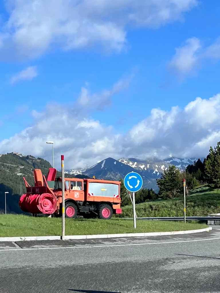 Eine große, orangefarbene Schneefräse steht als Dekoration auf einer Rasenfläche in einem Kreisverkehr vor schneebedeckten Bergen in Andorra.
