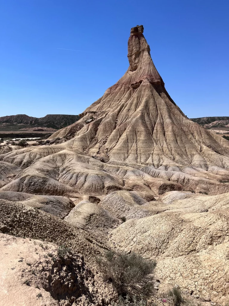 Ein Weitwinkel-Panoramablick über die erodierten, geschichteten Lehmhügel der Bardenas Reales. Im Zentrum steht die spitze, markante Felsformation. Der Himmel ist wolkenlos und blau.