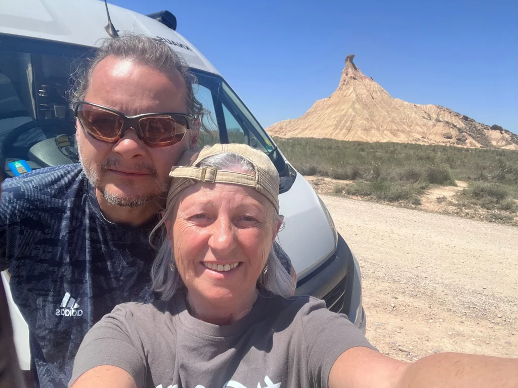 Ein Selfie-Foto von Ingo und mir (Kariin), beide lächelnd, vor unserem Kastenwagen. Ingo trägt eine Sonnenbrille, ich trage meine beige Kappe. Im Hintergrund ist die Wüstenlandschaft und eine Felsformation zu sehen.