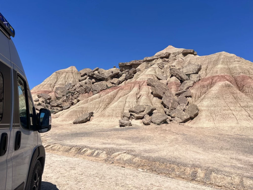 ine Seitenansicht des Kastenwagens auf einer Schotterstraße, der auf erodierte, geschichtete Hügel in der Halbwüste Bardenas Reales blickt. Die Felsen haben unterschiedliche Farben und sind mit großen Gesteinsbrocken bedeckt.