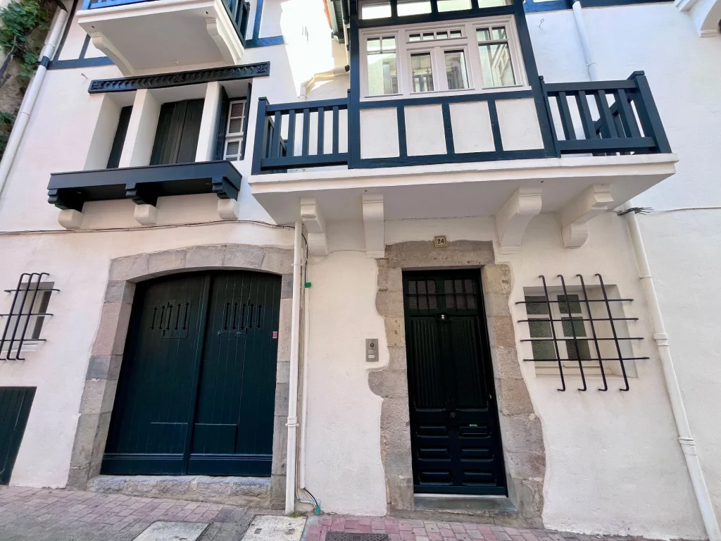 Traditionelle baskische Hausfassade in Weiß mit dunkelgrünen Holzbalkonen, Fensterläden und Steineinfassungen an den Türen.