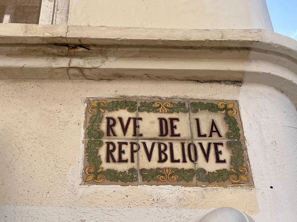 Ein kunstvolles, historisches Straßenschild aus verzierten Keramikfliesen mit der Aufschrift RVE DE LA REPVBLIOVE an einer Hauswand.