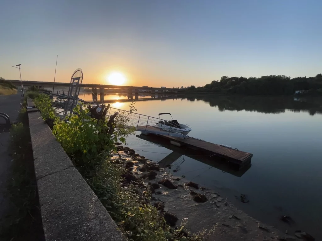 Ein kleiner Bootssteg am ruhigen Fluss Adour in Bayonne bei einem klaren, goldenen Sonnenuntergang mit einer Brücke im Hintergrund.